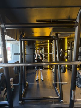 A person wearing a school uniform stands in the center of a dark, industrial-style gym with exposed black pipes and yellow lighting, posing for a mirror selfie.