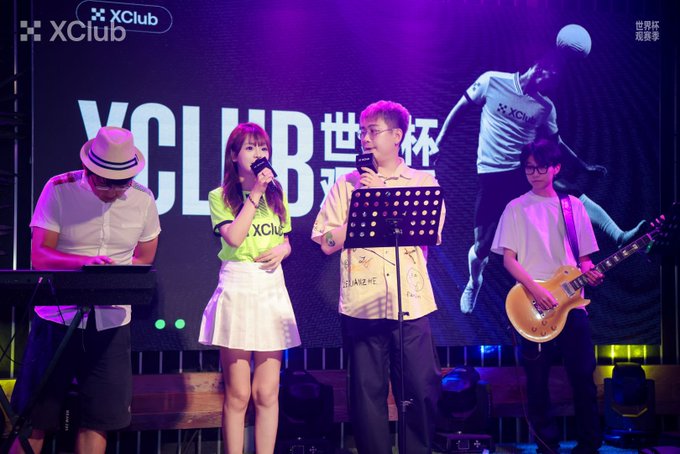 A live band performing on stage at 'XClub' World Cup event, with a female singer in a green XClub shirt, a male vocalist, a keyboardist in a hat, and a guitarist with a Les Paul guitar.