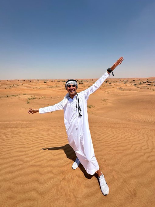 A person in a white robe and headscarf joyfully leaps with arms outstretched against a backdrop of vast orange sand dunes under a clear blue sky.