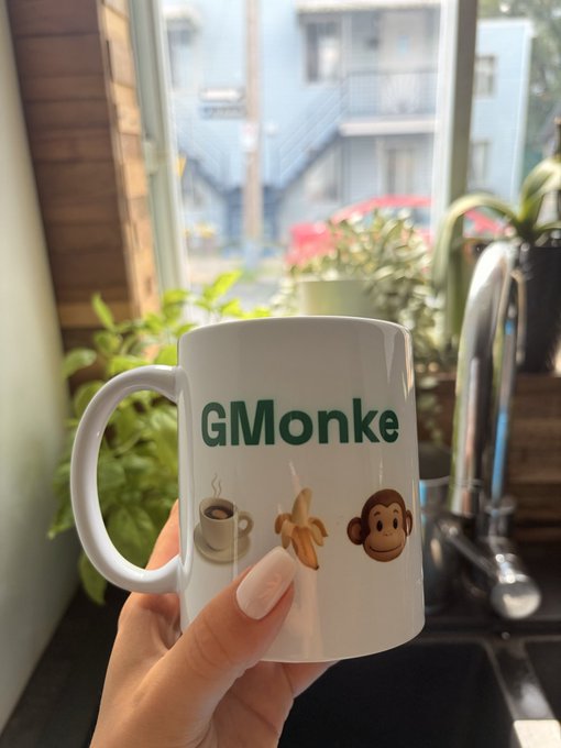 Photo of a hand holding a white coffee mug with 'GMonke' text and emojis of a coffee cup, banana, and monkey face, set against a kitchen window with plants and a view of buildings outside.