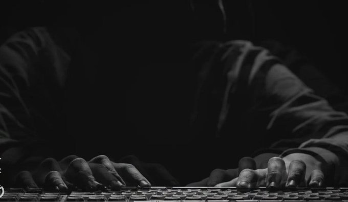A black and white close-up showing hands typing on a keyboard in a dark setting, with sleeves visible but the person's face obscured by shadow.