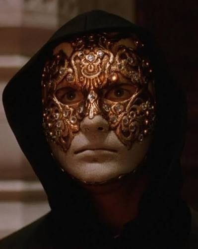 A close-up of a man wearing an ornate, jewel-encrusted golden half-mask that covers his eyes and nose, with a black hood framing his face against a blurred background.