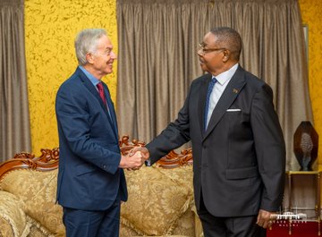 Two men in suits shaking hands in an ornately decorated room with gold curtains and furniture, appearing to be at a diplomatic or official meeting.