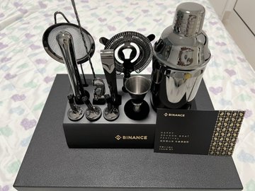 A sleek black Binance-branded gift box is displayed on a bed, containing a premium cocktail shaker set with chrome bar tools (strainers, jigger, stirrers), alongside a Binance gift card. The caption from the user thanks Binance for the high-end Dragon Boat Festival gift box, noting its impressive quality and thoughtful presentation.