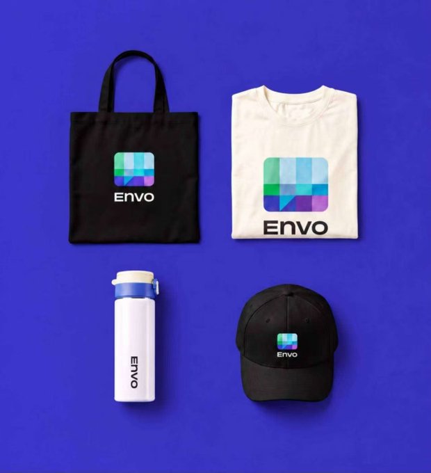 A branded merchandise display on a purple background featuring 'Envo' products: a black tote bag, a white t-shirt, a white water bottle, and a black cap, all displaying a pixelated blue-green-purple logo.