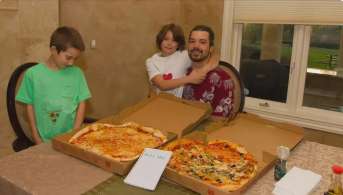 A family photo shows a man seated at a dining table with two young boys, with two open pizza boxes and a handwritten notepad on the table in front of them.