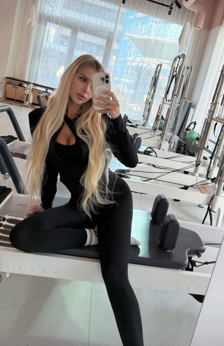 A woman with long blonde hair wearing a black workout outfit takes a mirror selfie in a Pilates/gym studio while sitting on Pilates equipment.