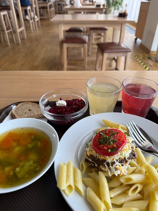 A photograph of a meal on a wooden table in a restaurant, featuring a bowl of vegetable soup, penne pasta with a meat patty topped with a tomato slice, a side of beet salad, bread, and two glasses of juice.