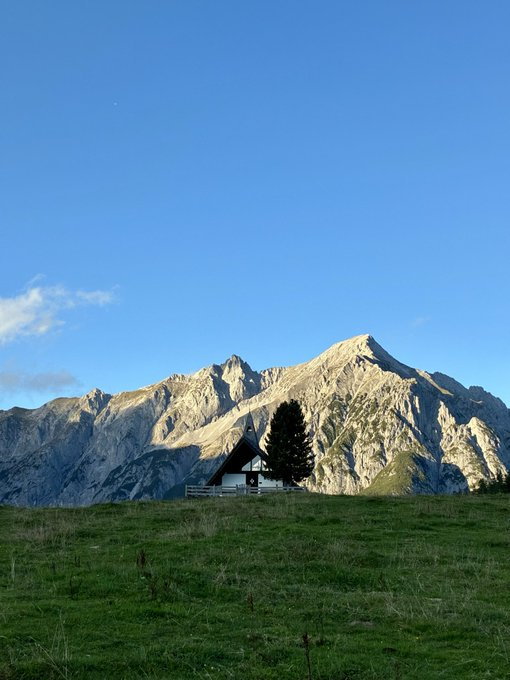 A serene landscape showing a small wooden cabin beside a pine tree on a green grassy meadow, with a large, rugged mountain range rising in the background under a clear blue sky.