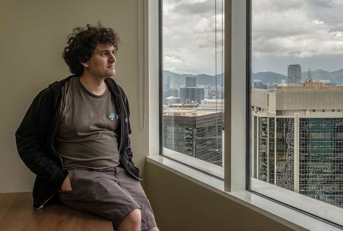 A man with curly hair sits on a windowsill, gazing out at a city skyline with mountains in the distance.