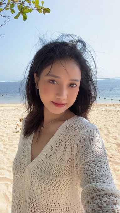 A young woman with dark hair wearing a cream-colored knit cardigan, taking a selfie on a sandy beach with the ocean behind her.