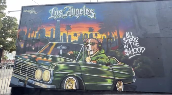 A colorful street mural in Los Angeles features a dog wearing headphones and a green hoodie driving a classic green convertible, with the text 'Los Angeles' and 'ALL $GOOD IN THE $HOOP' painted on the wall.