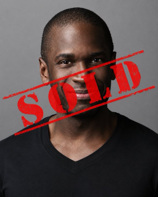 The image features a man wearing a black shirt, with red text that reads "SOLD" written above him. The man has a serious expression as he looks directly at the camera. The background of the image is gray, providing a neutral backdrop for the subject's attire and expression.