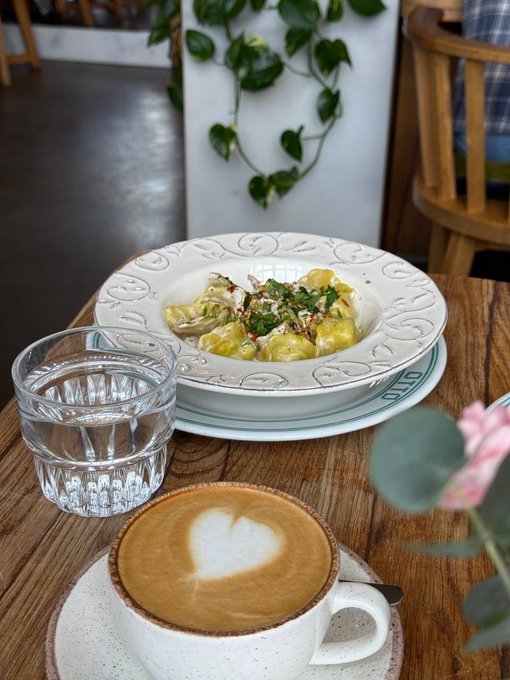 A meal at a café featuring a cappuccino with a heart-shaped latte art, a glass of water, and a plate of tortellini pasta garnished with herbs, accompanied by pink roses and trailing ivy.