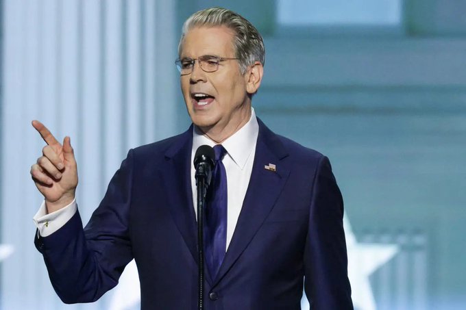 A man in a dark blue suit and tie with an American flag pin, wearing glasses, speaking at a microphone while pointing upward.