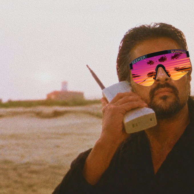Man with a beard wearing reflective pink 'SEEKER SUMMER' sunglasses and holding a large vintage brick-style cellphone to his ear, standing on a beach at sunset.