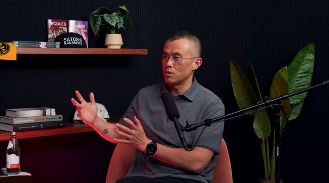 A man with glasses wearing a gray polo shirt speaking into a microphone in a studio setting, with a bookshelf behind him displaying titles like 'ÉCOLES', 'SATOSHI NAKAMOTO', and 'DeLorean Time'.
