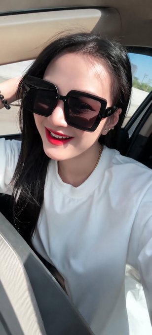 A woman wearing large black square sunglasses and a white t-shirt sits in a car, smiling brightly with red lipstick. Her confident, selfie-style pose from the driver's seat complements the post's theme of perseverance on 𝕏 (formerly Twitter), suggesting the resilience and self-assurance needed to keep climbing despite uncertainty.