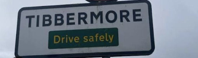 A white road sign with black text reading 'TIBBERMORE' and a smaller green sign below it displaying 'Drive safely' in yellow text.