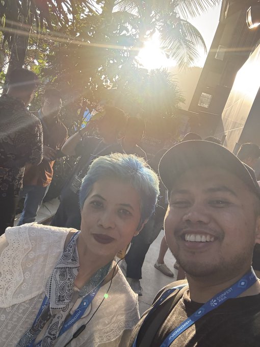 A selfie of two people at a SUNFEST event, with the man wearing a blue cap and a SUNFEST lanyard, and the woman with short blue hair wearing a lanyard over a patterned blouse, taken outdoors with bright sunlight and palm trees in the background.