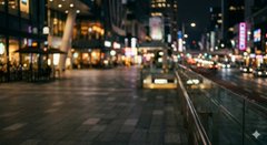 A blurry nighttime cityscape showing illuminated buildings, storefronts, and street lights along a pedestrian walkway.