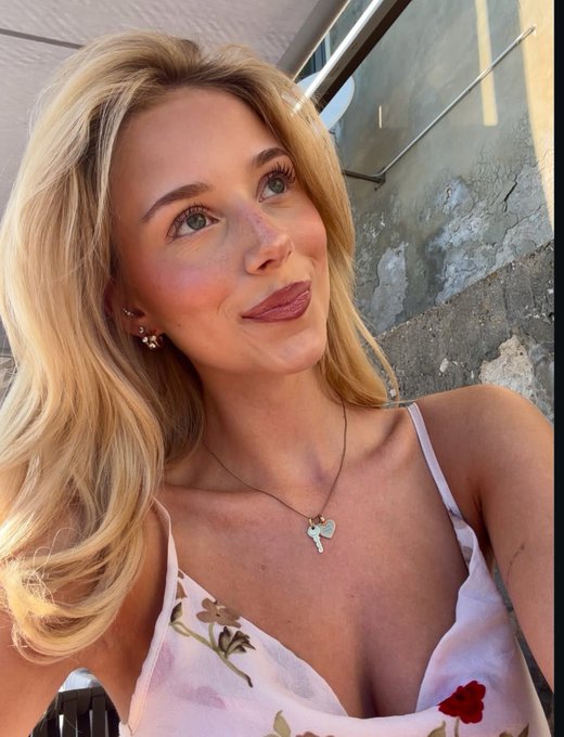 A blonde woman with blue eyes takes a selfie wearing a light pink floral dress and a silver heart-shaped pendant necklace.