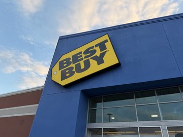 The image shows a blue building with a yellow Best Buy sign above it. The sign displays "Best Buy" in black letters against a white background. Below the sign, there are two windows of the store. A person can be seen walking by the store on the right side of the photo.