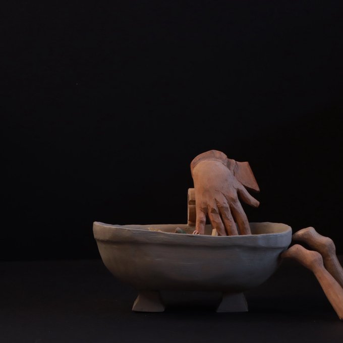 A surreal artistic composition featuring a terracotta hand emerging from a stone basin with a foot, set against a dark background.