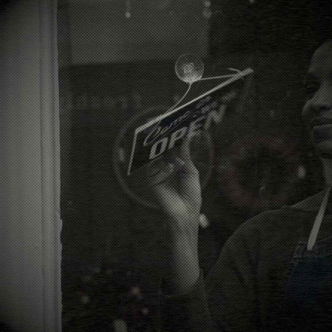 The image features a person holding an open sign that reads "Open". The scene takes place inside a building, possibly a store or office, with a window visible behind them. The background of the photo appears to be black and white, giving it a vintage feel. There is also a text overlay in the image which says "Solana will become the home of global finance. And global finance will run on smart accounts @altitude". This text seems to be related to the person holding the open sign or the building they are in.