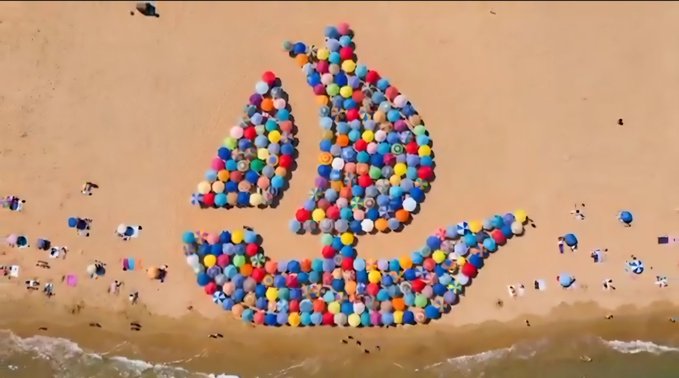 An aerial view shows a large crowd on a sandy beach arranging themselves with colorful umbrellas to form the shape of a sailboat, visually representing the "SEA" ticker and OpenSea brand mentioned in the celebratory Sunday post about the upcoming TGE.