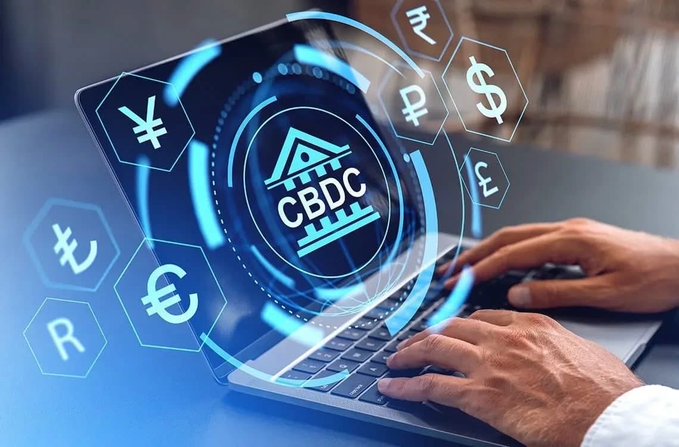 A conceptual image of hands typing on a laptop keyboard with a holographic 'CBDC' (Central Bank Digital Currency) emblem displayed above, surrounded by hexagonal icons featuring various currency symbols including $, €, ₹, ¥, ₽, and R$.