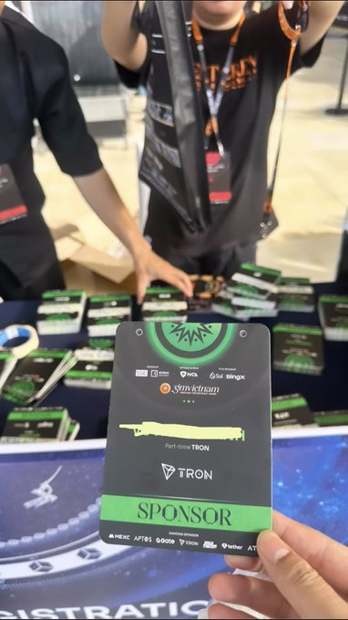 A hand holds a 'SPONSOR' pass for TRON at an event, with sponsors like BingX and HTX visible, while people stand behind a table with merchandise.