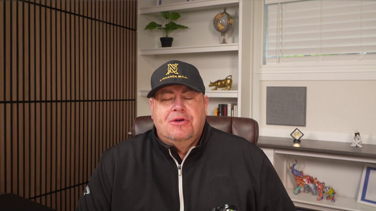 The image shows a man wearing a black jacket, sitting at a desk with his head down looking sad or thoughtful. The background of the image features a wall with a window that allows natural light into the room. There are also several books on the desk next to the man.