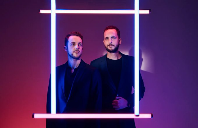 The image features two men standing next to each other, both wearing suits and ties. They are positioned within a frame that includes neon lights behind them, creating an interesting visual effect. The background color appears to be purple or pink, which complements the overall aesthetic of the scene. The image does not contain any text, but it seems to be related to a post on social media where two men in suits are being described as "immutable cuts" and "game dev teams."