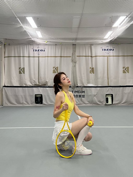 A woman in a yellow top and white skirt kneels on a tennis court holding a racket and ball, with banners reading "TIMDIM" and "币安网球" visible behind her. The image depicts the Binance tennis-themed rewards and fitness lifestyle she mentions, posing confidently before the start of her tennis lesson.
