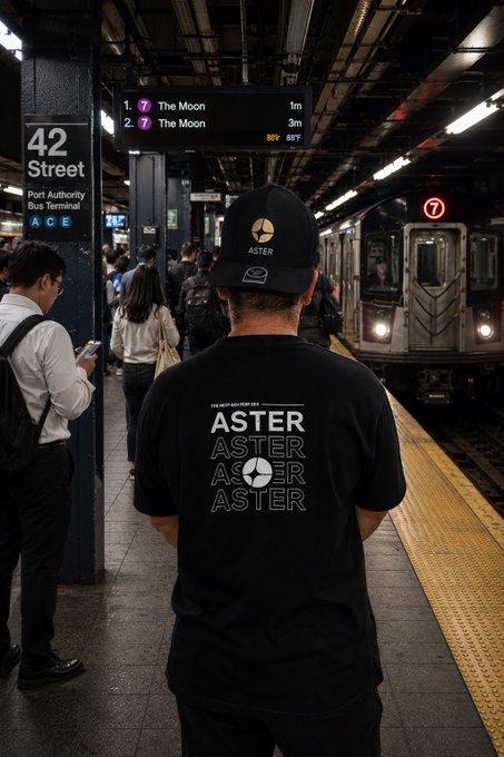 A person wearing a black 'ASTER' t-shirt and matching cap stands on the 42 Street subway platform in New York as a 7 train approaches, with a sign reading 'The Moon' displayed on the platform screen.