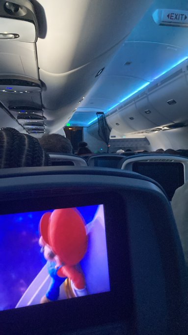 The interior of an airplane cabin lit with blue mood lighting, with a seatback screen showing an animated character resembling Mario.