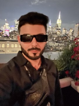 A man with dark hair, sunglasses, and a goatee wears a black shirt and shoulder bag, taking a selfie with a city skyline at night behind him.