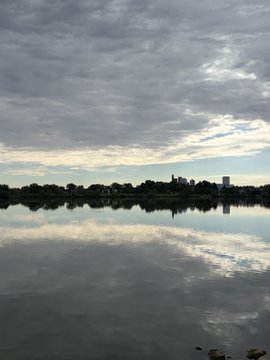 A serene landscape photograph of a calm lake reflecting a cloudy sky, with a dark silhouette of trees and a building on the distant horizon.