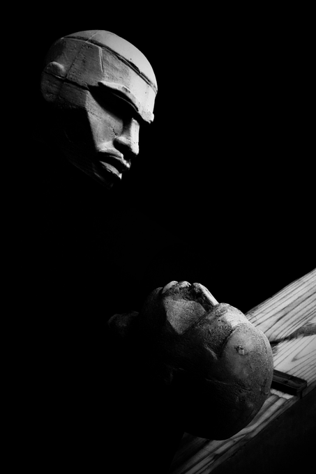 A dark, dramatic black and white photograph shows a sculpted head and a clenched fist emerging from deep shadow.