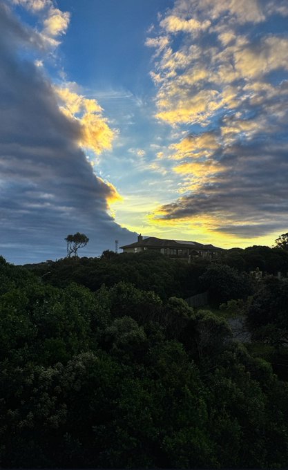 A scenic landscape photo showing a dramatic sunset sky with golden clouds above silhouetted trees and a house on a hill.