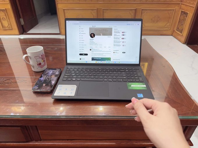 A workspace scene showing a laptop on a glass-topped wooden table displaying a webpage, alongside a smartphone, a Hello Kitty mug, and a person's hand resting on the table.