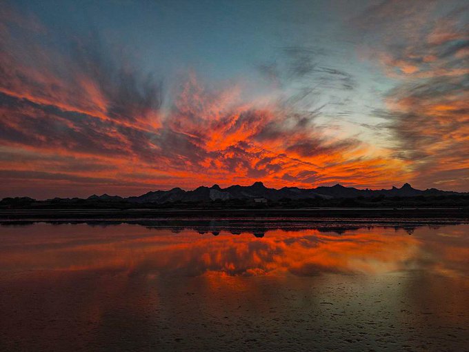 A vibrant sunset sky with dramatic red and orange clouds reflected in a calm body of water, silhouetting distant mountains on the horizon.