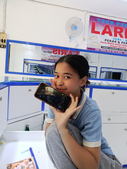 A young woman smiling while holding up a smartphone to her ear in what appears to be a shop or stall, with Indonesian text visible on banners reading 'TOKO PER... LARIS' and 'ARIS' in the background.