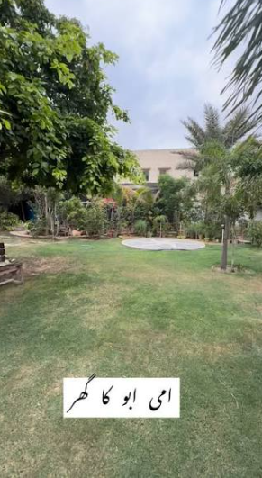 A grassy backyard garden with trees and a building in the background, featuring Urdu text 'اسی ابو کا گھر' (This is Abu's house) at the bottom.