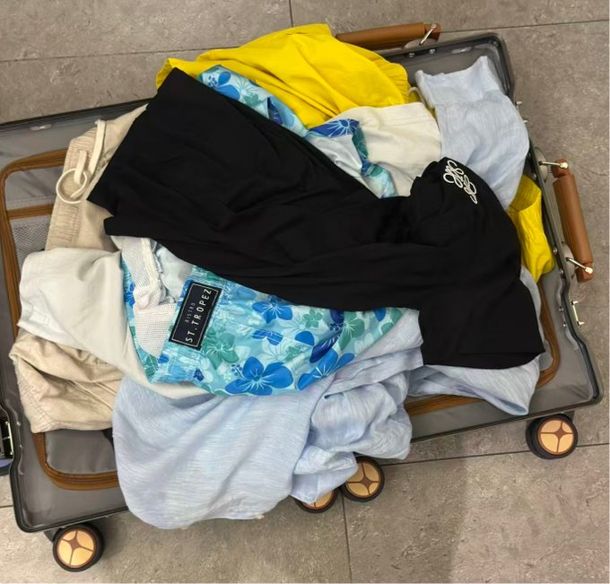 An open suitcase lies on a tiled floor, filled with folded clothing in various colors including yellow, black, white, and a blue floral pattern.