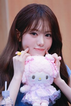 A young woman with long brown hair and bangs poses with her hands near her face while holding a pink and purple Hello Kitty plush toy.