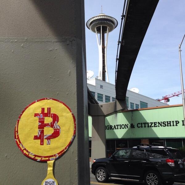 A street view featuring a yellow and red Bitcoin sign attached to a pole, with the Space Needle and a building labeled 'MIGRATION & CITIZENSHIP' in the background.