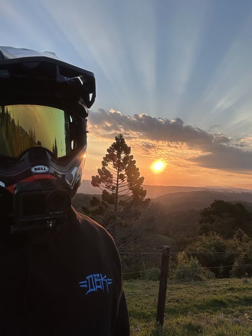 A person wearing a Bell helmet with attached action camera poses at sunrise over a hilly landscape, with "ZINK" branding on their hoodie, fitting the "GM, have a blessed new week" theme by capturing the start of a new day.