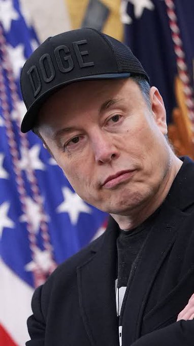 Elon Musk wears a black "DOGE" cap while standing in front of an American flag, appearing to subtly hint at his support for the Dogecoin meme coin. The image aligns with the post's suggestion that Musk often drops subtle clues about the cryptocurrency he promotes.